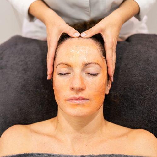 Crop cosmetologist doing facial massage of female customer lying on towel with closed eyes and applying cleansing mask on face during skin care routine in beauty center